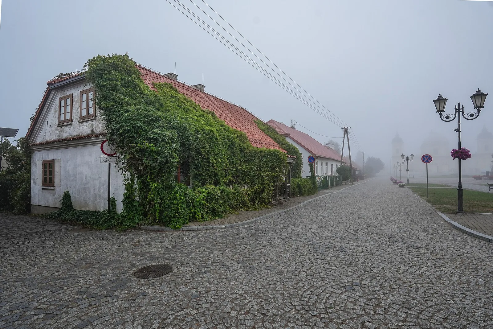 Tykocin , a historic house on a cobblestone street near the market square, partly covered with ivy, in fog. The photograph shows a white-plastered building with a red ceramic-tile